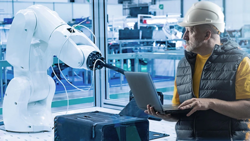 Image of a man in a hardhat holding a laptop and looking at an industrial automation arm